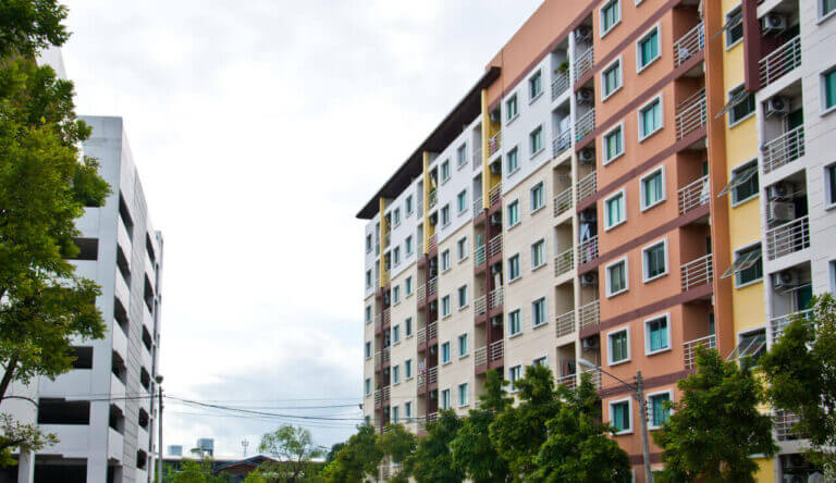 Two multi-story apartment buildings are seen, one white on the left and one with pastel-colored sections on the right. Both have multiple windows. Trees and a few power lines are visible in the foreground, while a locksmith's van is parked nearby under the cloudy sky in the background.
