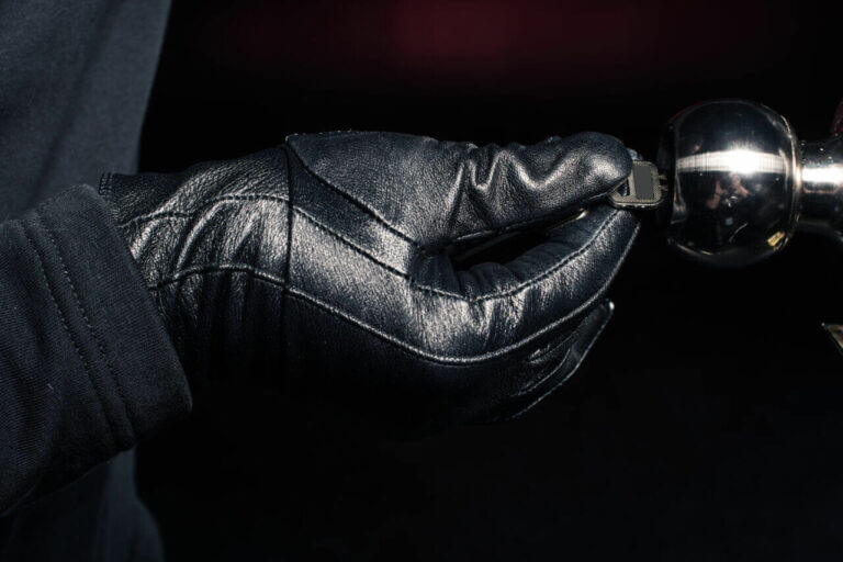 A person wearing black leather gloves, resembling a skilled locksmith, is inserting a key into a silver lock. The close-up image highlights the gloved hand and the lock, set against a dark, blurred background.
