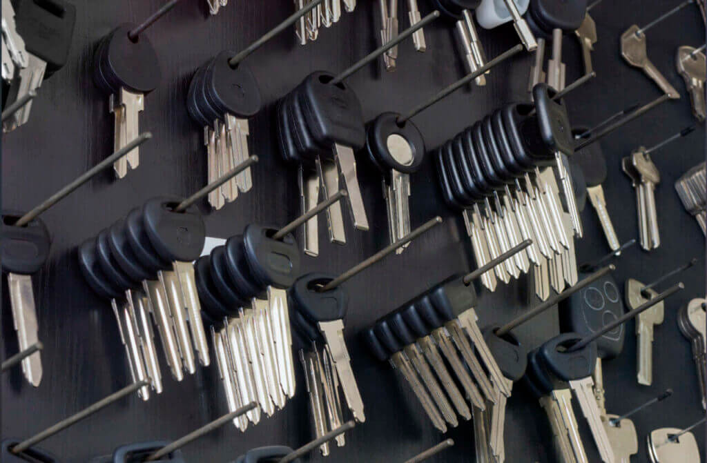 A wall-mounted board, reminiscent of a locksmith’s workspace, showcases numerous sets of keys organized on hooks. Each set, featuring black plastic heads, is arranged in rows depicting various types. Some keys have distinct shapes for different locks, with a few individual ones scattered around.