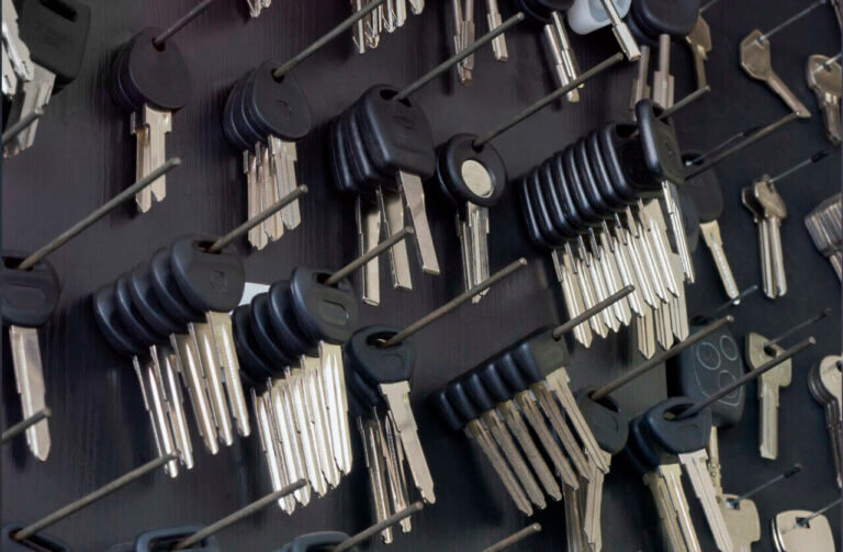 A wall-mounted board, reminiscent of a locksmith’s workspace, showcases numerous sets of keys organized on hooks. Each set, featuring black plastic heads, is arranged in rows depicting various types. Some keys have distinct shapes for different locks, with a few individual ones scattered around.