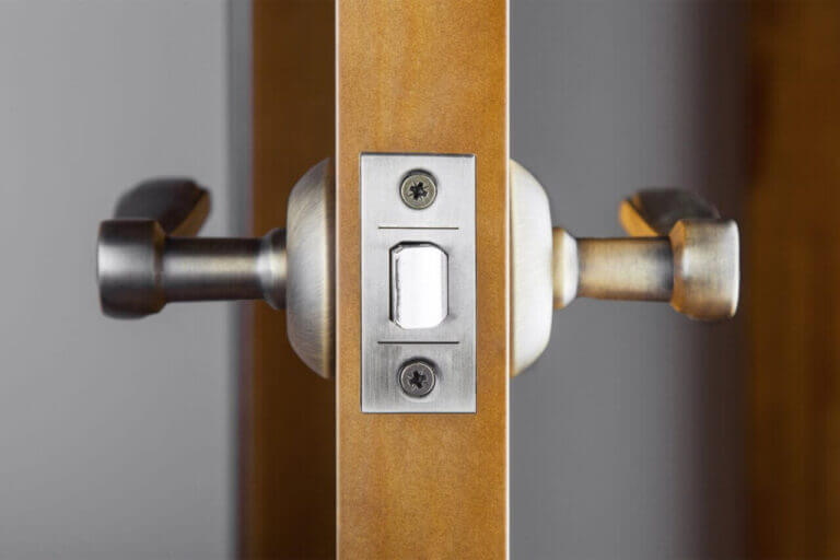 Close-up of a door latch mechanism installed on a wooden door. The latch is made of metal with a rectangular faceplate secured by two screws. A locksmith would appreciate the sturdy, metallic and curved door handles on both sides. The background appears blurred.