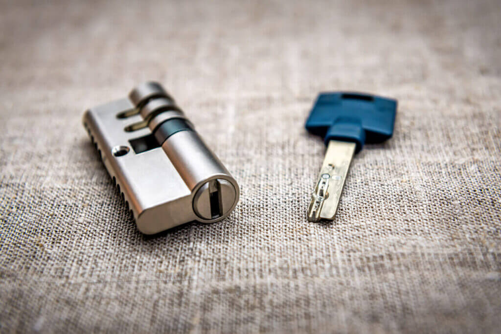 A locksmith's metallic cylinder lock mechanism and a silver key with a blue plastic grip are placed on a textured beige fabric surface. The lock is on the left, and the key is on the right side of the image.