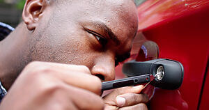 Close-up of a locksmith using a tool to unlock a car door. The individual is focused, with one hand holding the tool and the other stabilizing the car’s door handle. The car is red and the scene is set outdoors.