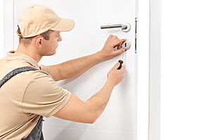 A locksmith wearing a beige cap and uniform is meticulously working on a white door, skillfully using tools to adjust the lock mechanism. The door features a modern handle, and the focused locksmith appears dedicated to ensuring everything is in perfect order.