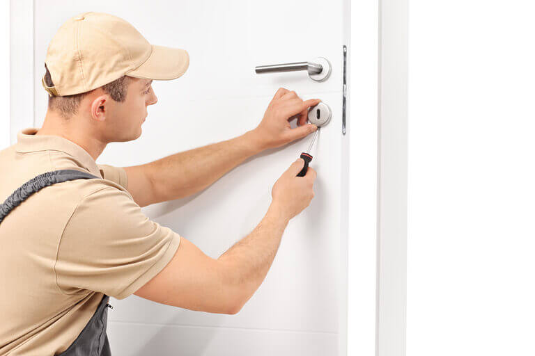 A locksmith wearing a beige cap and uniform is meticulously working on a white door, skillfully using tools to adjust the lock mechanism. The door features a modern handle, and the focused locksmith appears dedicated to ensuring everything is in perfect order.