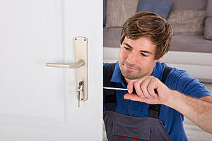 A locksmith in a blue shirt and grey overalls is using a screwdriver to adjust the lock of an interior door. The door is white and slightly open, with a silver handle and key inserted in the lock. The background shows a couch and a beige wall.