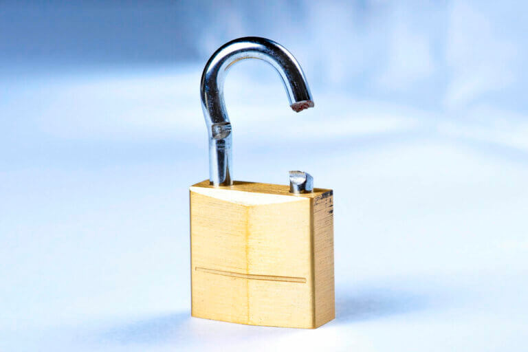 A close-up image of a broken padlock. The padlock, clearly illustrating why a skilled locksmith is essential, is made of brass with a silver shackle, which is bent and snapped on one side, rendering it non-functional. The background is out of focus, making the padlock the focal point of the image.