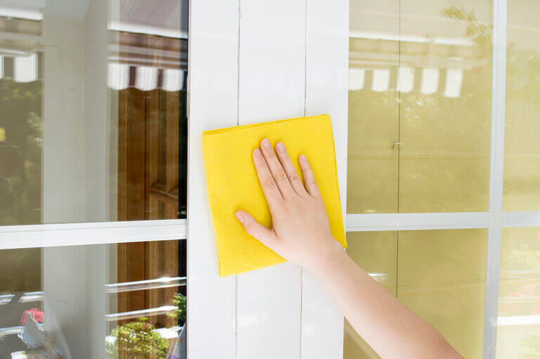 A hand holding a yellow cloth is seen cleaning a glass surface of a window with white frames, as though meticulously ensuring every detail is spotless—much like a locksmith perfecting his craft. The background shows an outdoor view with greenery and a striped awning.