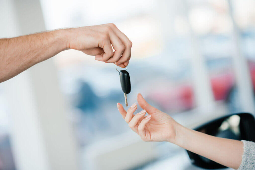 A close-up of one hand passing a car key to another hand in a smooth transaction. The background is blurred, showing an indoor setting with glass windows and hints of red and blue objects, as if in a locksmith's shop.