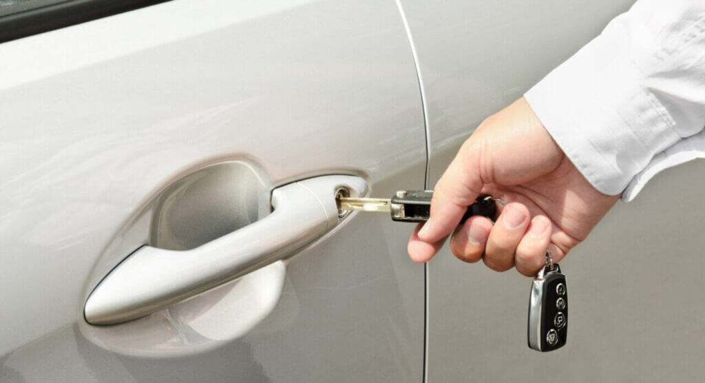 A hand holding a car key is inserting it into the keyhole of a silver car door, resembling the precision of a locksmith. The person is wearing a white long-sleeve shirt, and a key fob with buttons is attached to the key. The close-up shows only the hand and part of the car door.
