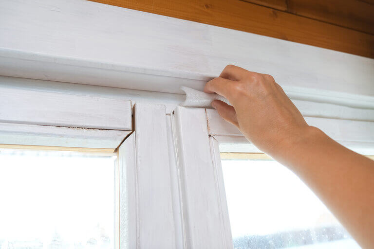 A hand is seen applying weatherstripping foam tape to a window frame, meticulously improving insulation. The white-painted frame, bordered by wooden trim, gleams in the sunlight. This home interior project might even impress a locksmith with its attention to detail and security.