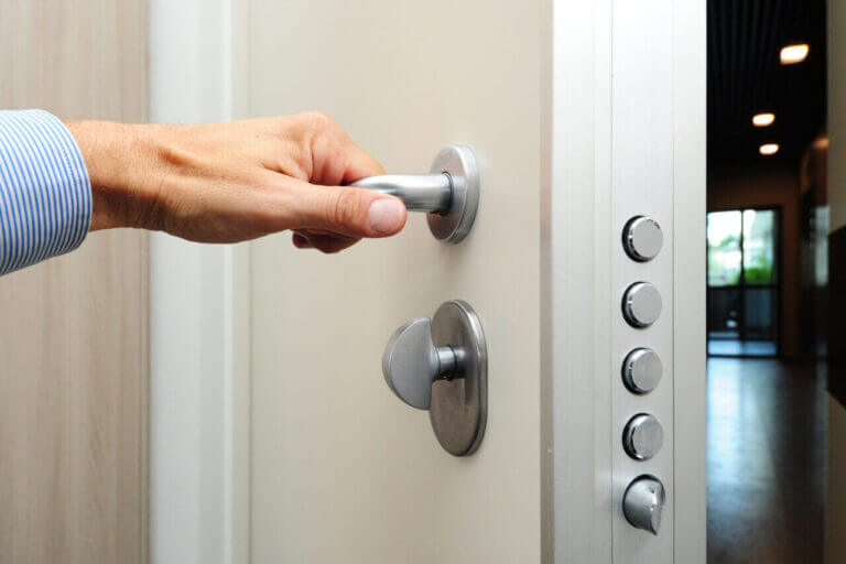 A person with a blue-striped shirt is turning the metal door handle to open a white door, fitted with a series of cylindrical locks, reminiscent of what a locksmith might install. The surrounding area appears to be indoors.