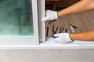 Close-up of hands wearing white gloves with green trim, installing a sliding glass door. Various tools, typically used by a locksmith, including screwdrivers, pliers, and a caulking gun, are scattered on the wooden floor in the background.