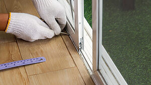 A locksmith wearing white gloves is carefully inserting a small tool into the bottom track of a sliding metal screen door. The door is positioned on a wooden floor, with a ruler lying beside it. Outside, a patch of green artificial grass is visible.