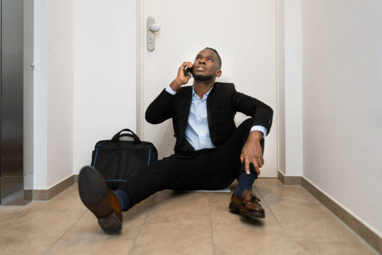 A man in a black suit sits on the floor in a narrow hallway, leaning against a door while talking on his phone. Beside him is a black bag that hints he might be awaiting a locksmith. The floor is tiled, and the walls are pristine white as he glances upward.