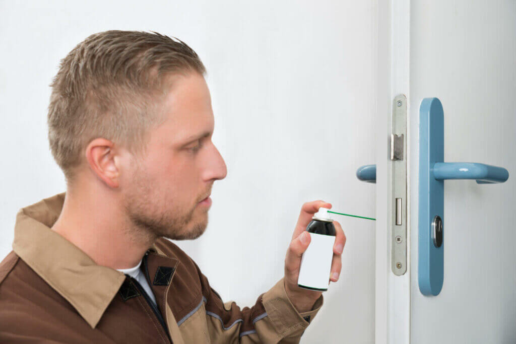 A locksmith is using a lubricant spray on a door lock. He is wearing a brown and beige jacket and focusing on applying the lubricant to ensure the lock functions smoothly. The door handle and lock mechanism are visible in the close-up image.