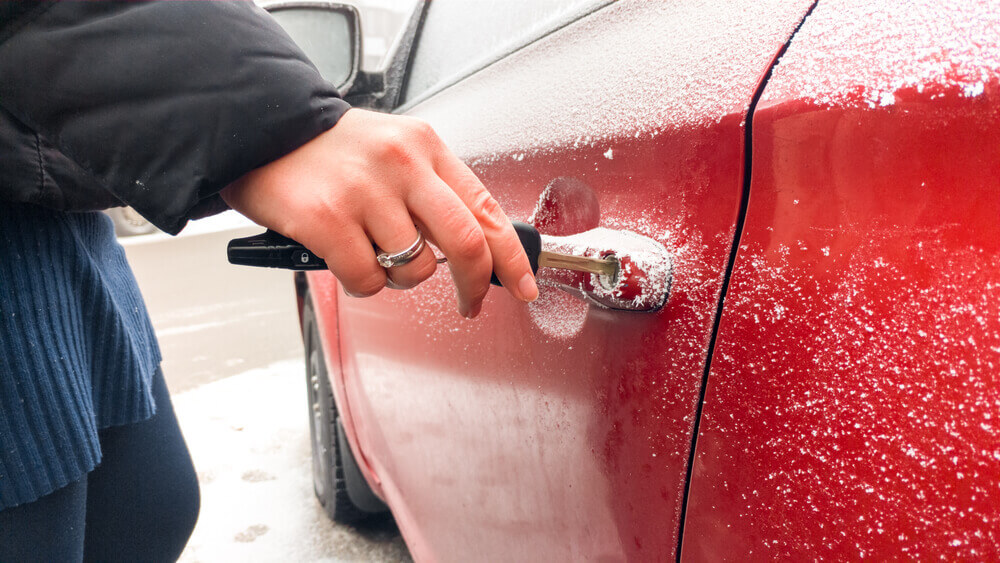 A locksmith is unlocking a frosty red car door with a key. Their hand, wearing a silver ring and holding a black car remote, meticulously works despite the light layer of ice and snow covering the car.