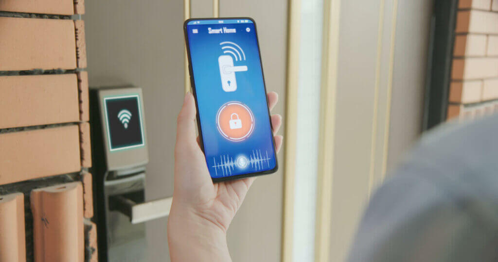 A person holding a smartphone displaying a smart home app with a lock icon. The phone is near a smart lock mounted on a brick wall, highlighting the seamless wireless connection—it's as if they have their own digital locksmith at their fingertips.