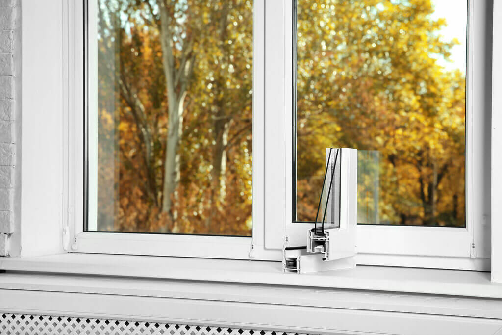 A close-up of a white window frame with a cross-section model of the frame sitting on the window sill. A locksmith's tools rest nearby, hinting at recent work. Outside the window, there are trees with autumn foliage.