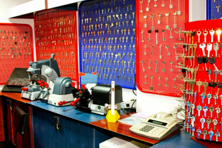A well-organized locksmith key cutting station is equipped with various key-cutting machines, a phone, and racks full of blank keys of different shapes and sizes on red and blue boards. The countertop is wood, and the cabinets below are blue.