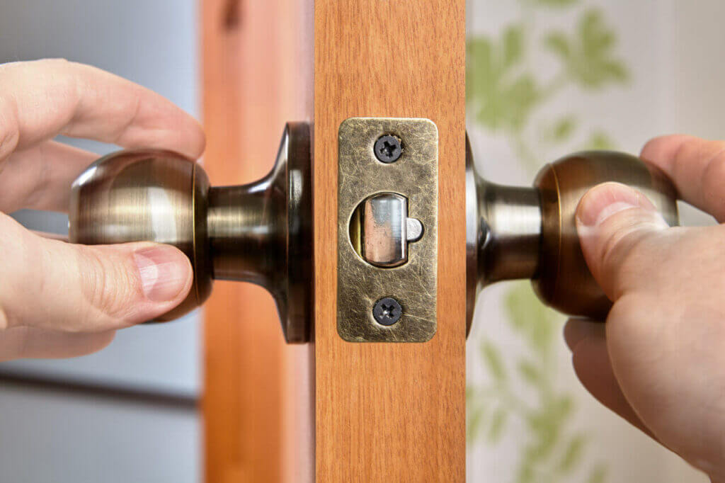 Close-up of a locksmith's hands installing a doorknob on a wooden door. The door is partially ajar, revealing the latch mechanism. The metallic hardware is secured with two screws, and the background includes a wall with leafy green patterns.