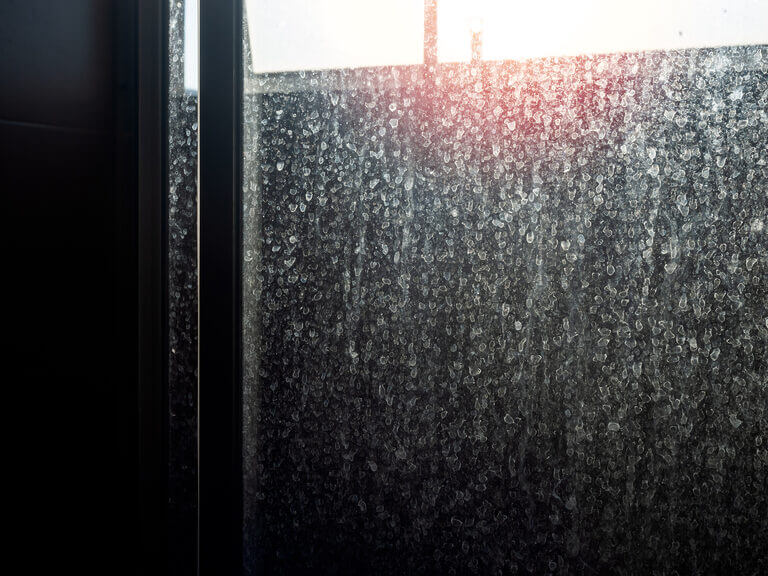 A close-up view of a window with numerous water droplets on the glass. Sunlight is faintly shining through the top portion, creating a slight red glow, while the background remains dark and blurry. A locksmith's reflection subtly intermingles with the droplets, adding an element of mystery.