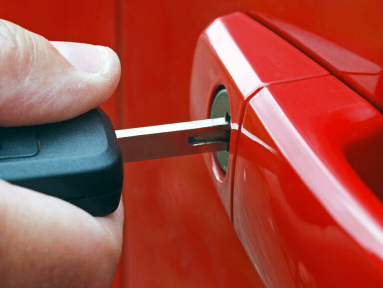 A close-up image of a hand inserting a key into the lock of a red car door, reminiscent of a locksmith at work. The key is partially inside the lock, and the keychain is visible in the background. The car's red door handle is also shown in the image.