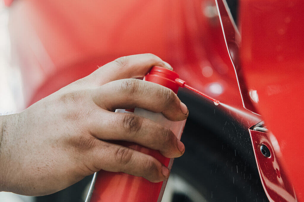 A close-up of a hand spraying lubricant from a red and white can onto the hinge area of a red car door. The focus is on the spraying action and the can, reminiscent of how a locksmith might ensure smooth operation, with a blurred background of the vehicle.