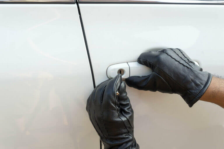 A person wearing black leather gloves is attempting to unlock a white car using a locksmith's tool near the door handle. The image focuses on the gloved hands and the car door.