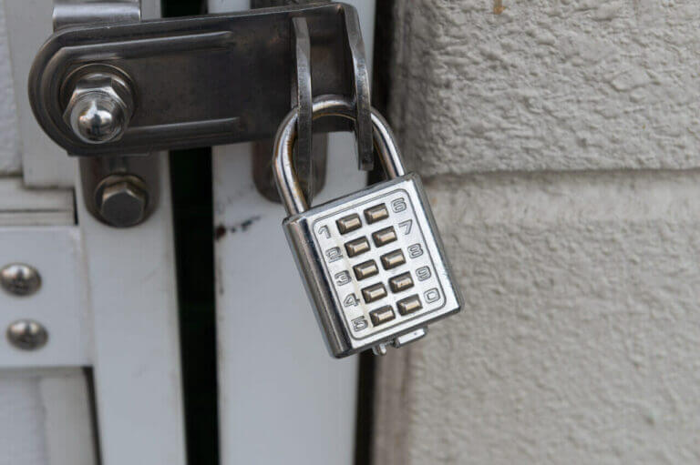 A closed combination padlock, securely fastened to a latch on a white, metal door, invites the skill of any seasoned locksmith. The lock's silver body is rectangular with a grid of numbered buttons for the combination. The surrounding wall appears textured and painted in a light color.