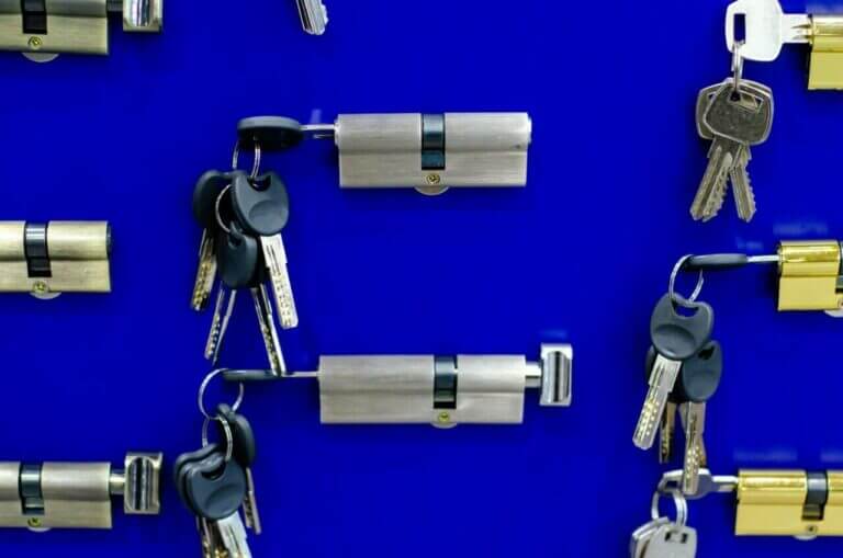 A locksmith's display of various cylindrical door lock mechanisms and sets of keys arranged against a bright blue background. Each lock has one or more keys attached, with the components neatly organized in a pattern.