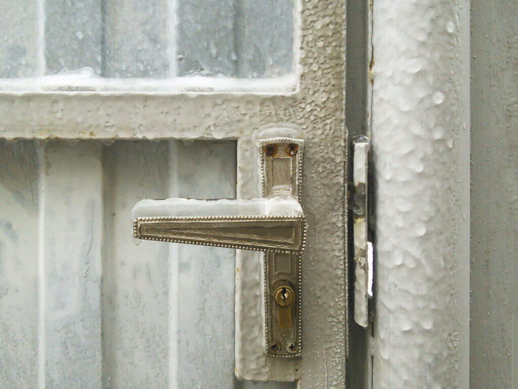 A metal door handle frozen over with a thick layer of ice is attached to a frosty door. The door and surrounding metal surfaces are covered in frost and ice, signaling the need for a skilled locksmith to navigate these extremely cold weather conditions.