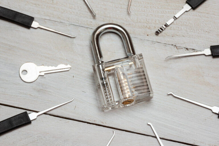 A transparent padlock is placed at the center of a wooden surface, surrounded by various lock picking tools and a key, reminiscent of a scene from a locksmith's workshop.