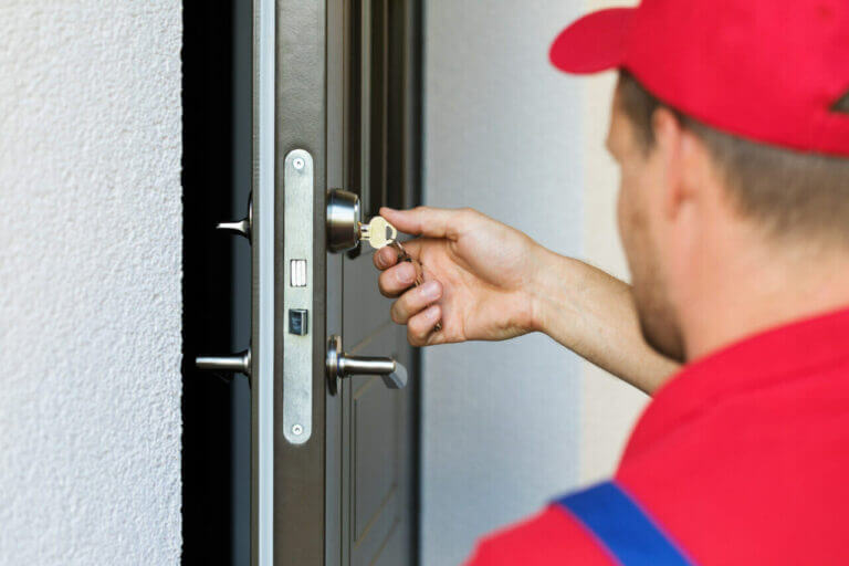 A locksmith in a red cap and shirt is seen from behind as they insert a key into a door lock. The individual is holding the key with their right hand, while the door features a modern handle and lock mechanism. The scene appears to be outdoors.