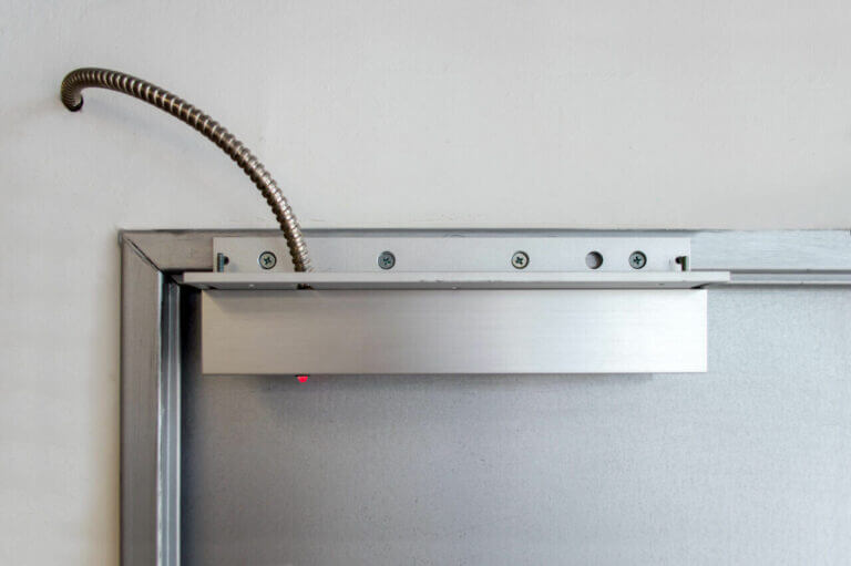 A close-up image of an automatic door closer mechanism installed by a locksmith at the top of a door frame. The device is metallic with a visible flexible conduit connected to it. Several screws secure the mechanism in place, and a red light indicator is visible.