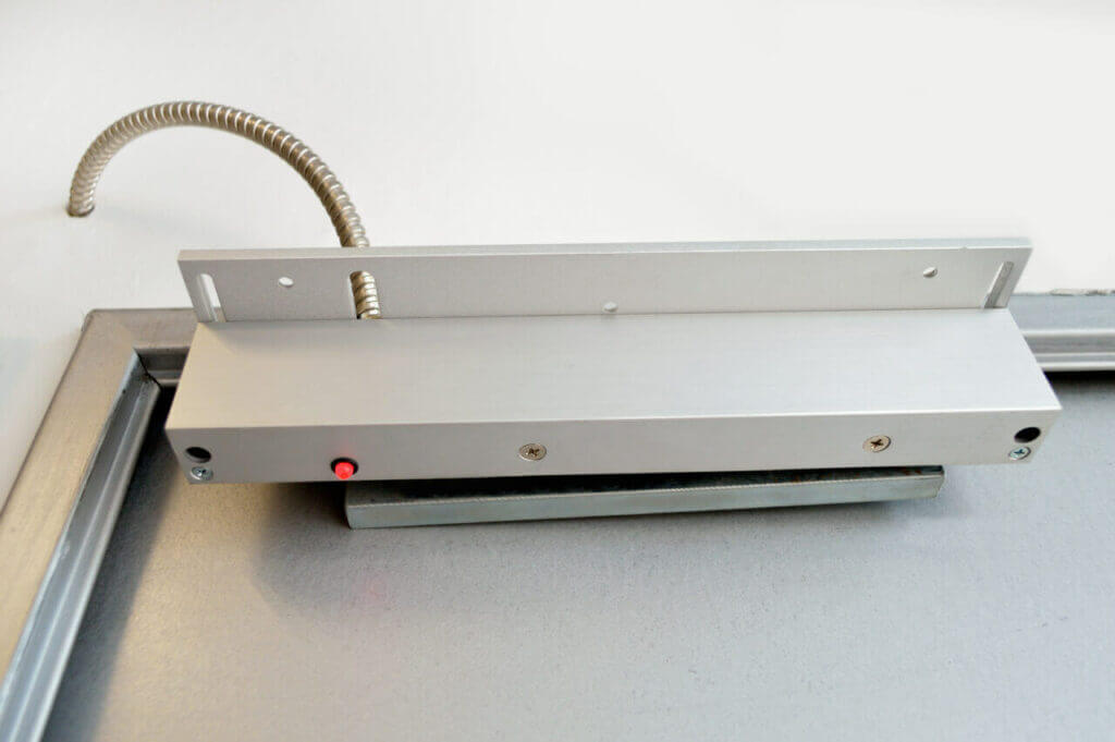 A silver electromagnetic door lock with a visible coil cord at the top is mounted onto a metal door frame. There is a red indicator light on the lock. This setup, likely installed by a skilled locksmith, appears to be part of a security system.