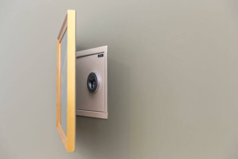 A hidden safe embedded in a wall, partially concealed by a picture frame. The frame is swung open, revealing the safe's door and combination lock behind it. The wall, painted a muted green color, might require the expertise of a skilled locksmith to access its secured contents.