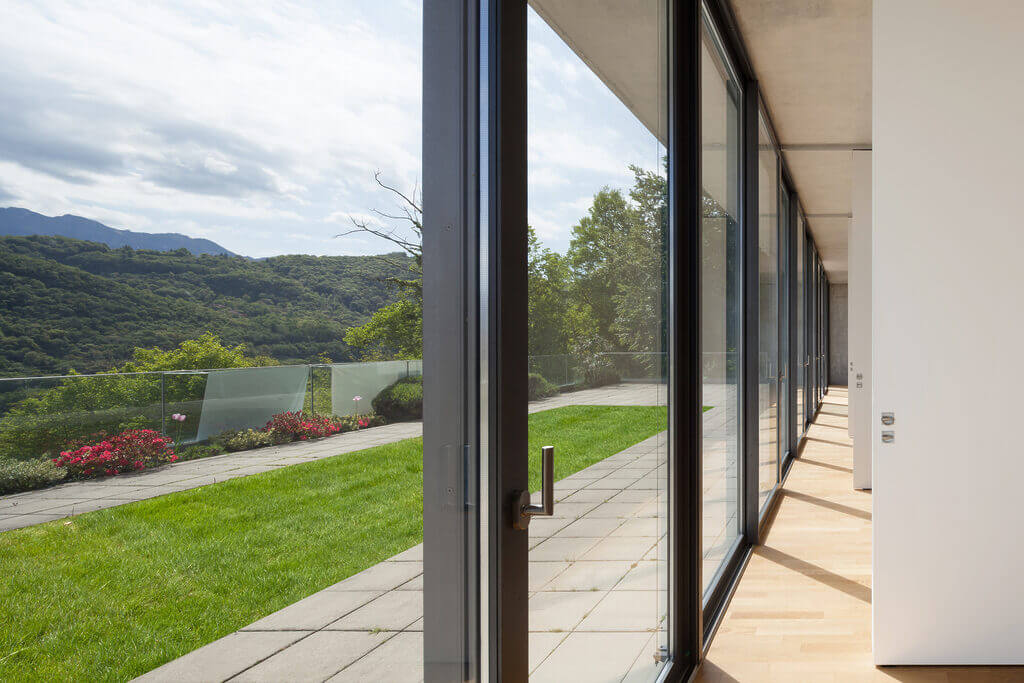 A corridor with large floor-to-ceiling glass windows on one side offers a view of a grassy patio, decorative plants, and distant green hills. The corridor floor is wooden, and a narrow concrete walkway runs parallel to the windows outside, adding charm akin to a locksmith crafting intricate details.