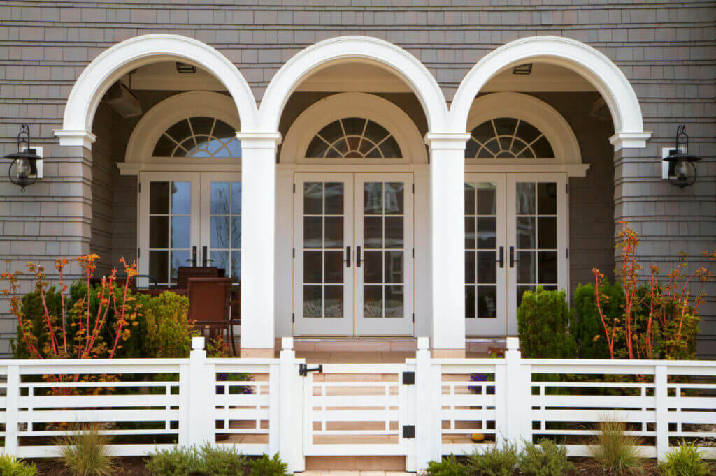 A house exterior featuring three identical white arched doorways with glass panes. Each doorway is flanked by two shuttered windows, and there is a small fenced front garden with shrubs and plants. A white picket gate and fence in the foreground completes this charming, secure abode—perfect for visiting a locksmith.