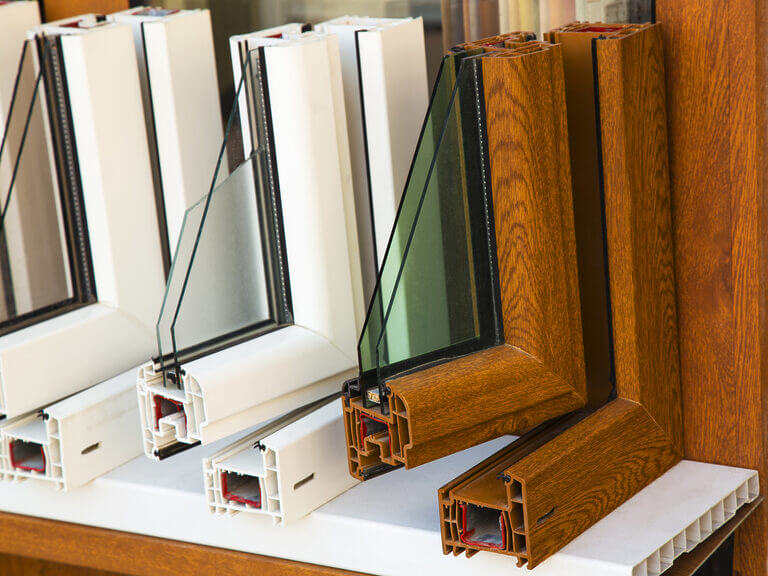 Display of cross-sections of window frames with double glazing. The white frames are on the left, and the wood-finish frames are on the right. All frames, reminiscent of a locksmith's precision, show the internal structure and insulation details. They are placed on a white surface.