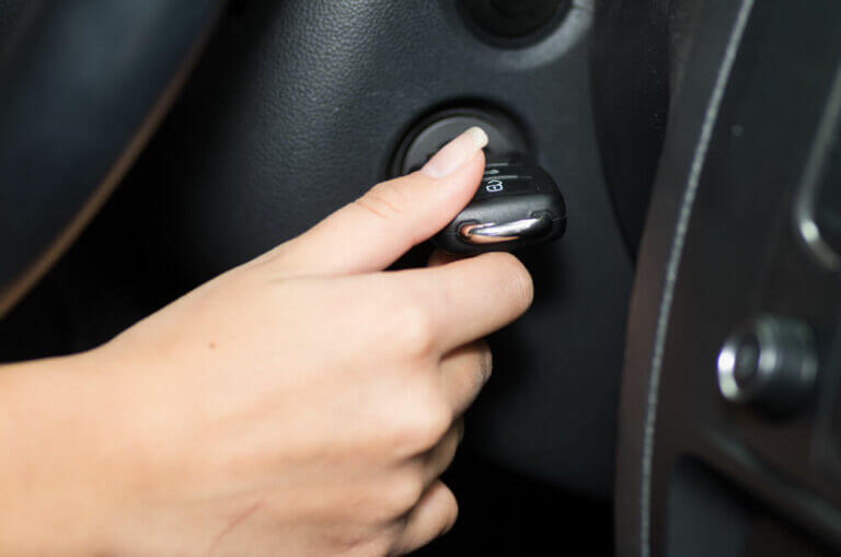 A close-up of a person's hand, reminiscent of a skilled locksmith’s precision, turning a car key in the ignition. The dashboard and steering wheel are partially visible, capturing the exact moment of starting the vehicle.