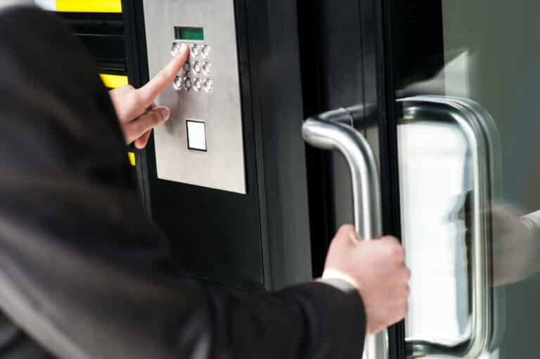A locksmith in a suit enters a code on a keypad next to a door with a metal handle. The person's left hand holds the handle while their right hand presses the keypad buttons.