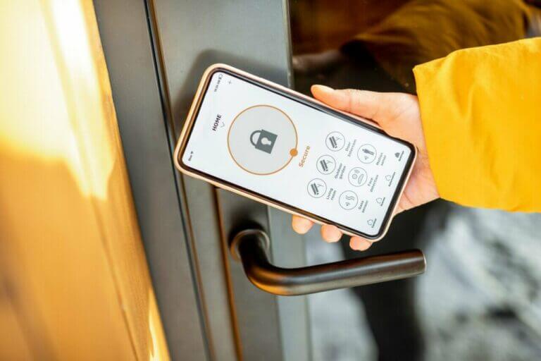 A person wearing a yellow jacket is using a smartphone to unlock a door with a smart lock application. The phone screen displays the app’s interface with a large lock icon and several options below it, making them look like a digital locksmith. The door has a black, modern handle.