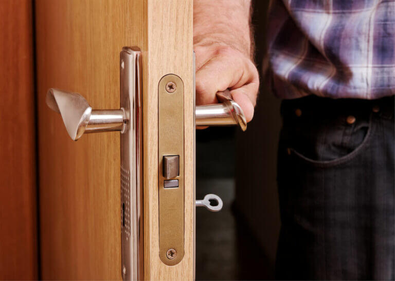 A person wearing a plaid shirt is opening a wooden door using a cylindrical silver door handle. The door, expertly installed by a locksmith, has a brass lock mechanism with a small key inserted into the keyhole. Only the person's hand and partial arm are visible.