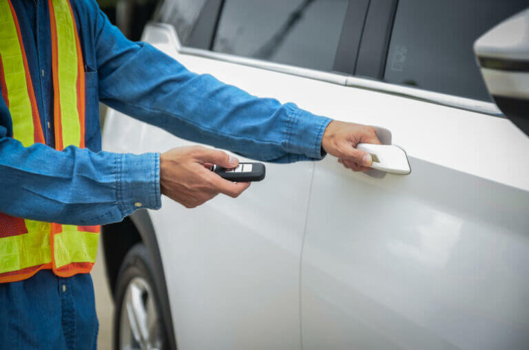 A locksmith wearing a high-visibility vest and a blue long-sleeved shirt is seen unlocking a white vehicle's door with a remote key. Only the person's hands and part of the upper body are visible. The car is parked outdoors.