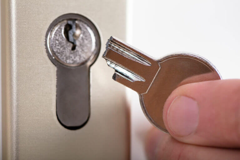 A close-up image of a person's hand holding a broken key in front of a door lock. The metal key is broken at the end, and the lock is seen against a plain background, just waiting for the skilled touch of a locksmith.