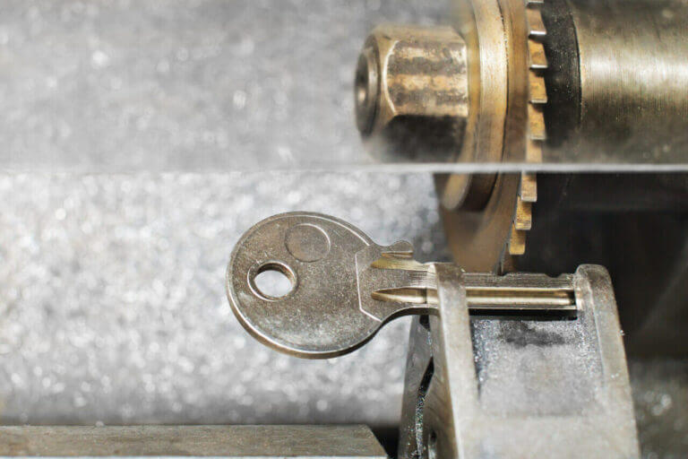 Close-up of a key being cut with a locksmith's precision using a key cutting machine. The machine's brass and steel components are visible, with the key clamped in place. The image focuses on the intricate cutting process.