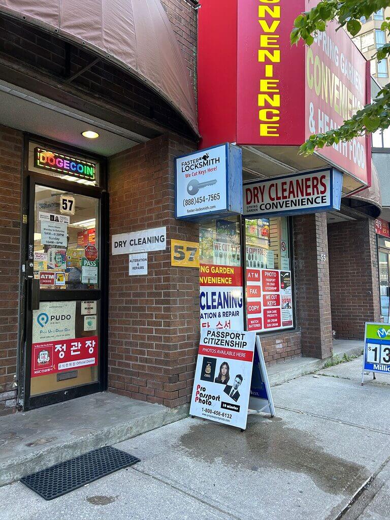 Street view of a storefront with multiple businesses. Signs indicate a dry cleaner, Faster Locksmith, and convenience store. An advertisement stand outside promotes passport photo services and phone repair. The entrance doorway displays various stickers and signs.