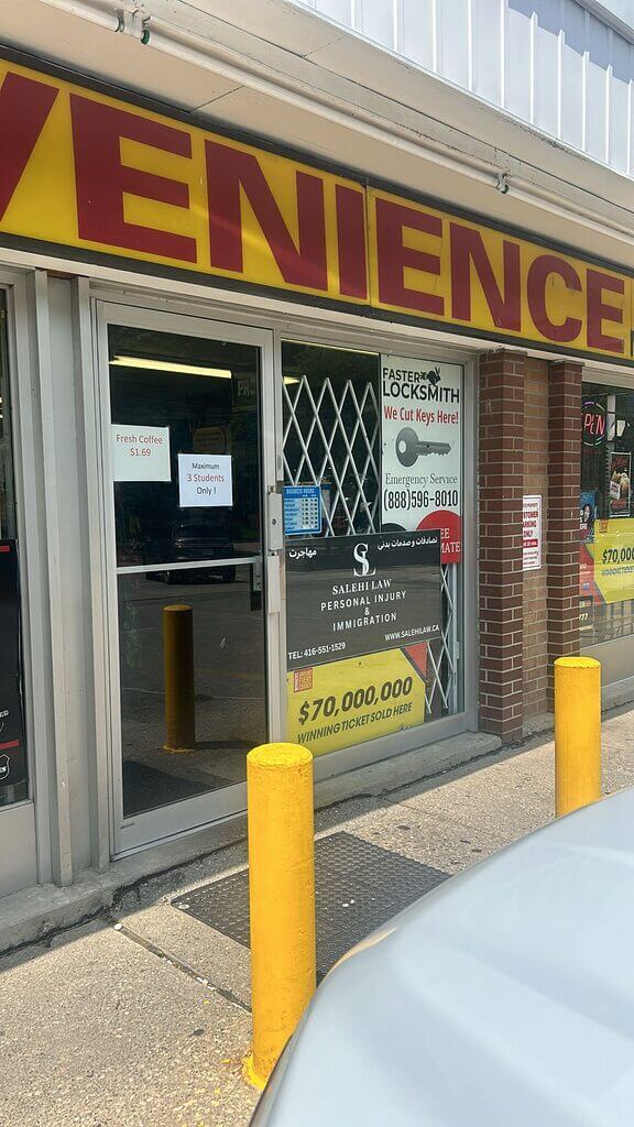 A convenience store storefront displays various signs, including “Faster Locksmith” with contact info. The door is reinforced with bars over the glass and yellow bollards stand out front.