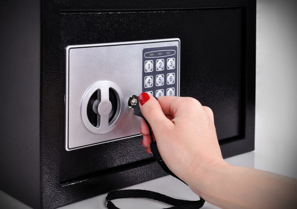 A skilled locksmith's hand with red nail polish is inserting a key into the lock of a black safe featuring a digital keypad. The safe boasts both a combination lock and key entry for enhanced security.